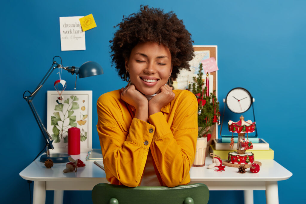 pleasant looking woman sits with closed eyes, toothy smile on chair, has good dreams, enjoys domestic atmosphere, poses against white desk in study room dressed in yellow jacket keeps hands under chin