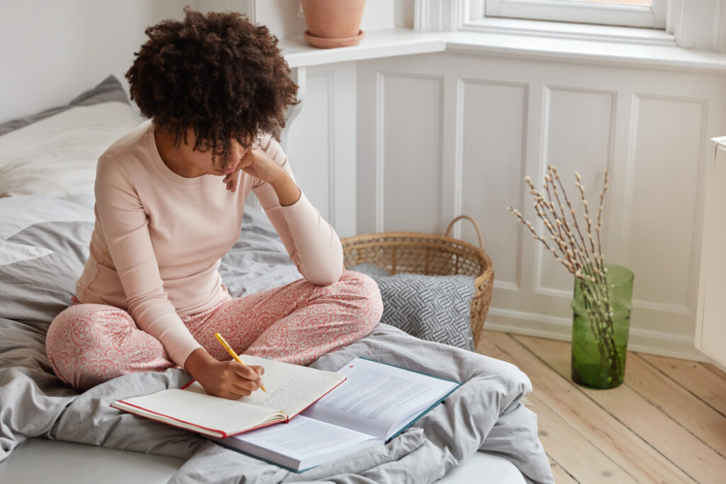 top view of busy dark skinned woman writes down records in notebook from book, has serious expression, poses on bed, enjoys comfort in bedroom, prepares for examination at college or university
